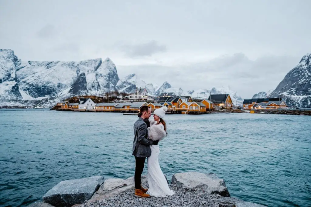 Lofoten Norway elopement couple stands on a dock with fjords, yellow fishing huts and snow-capped mountains behind