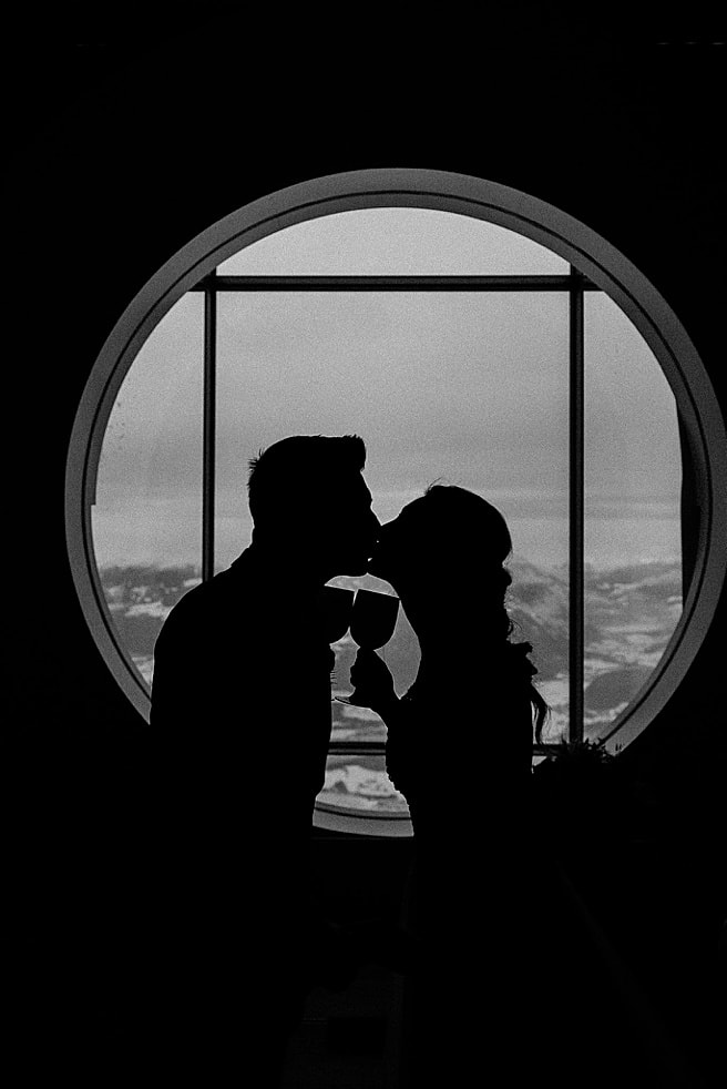 black and white image of a bride and groom kissing in front of a round window holding wine glasses.