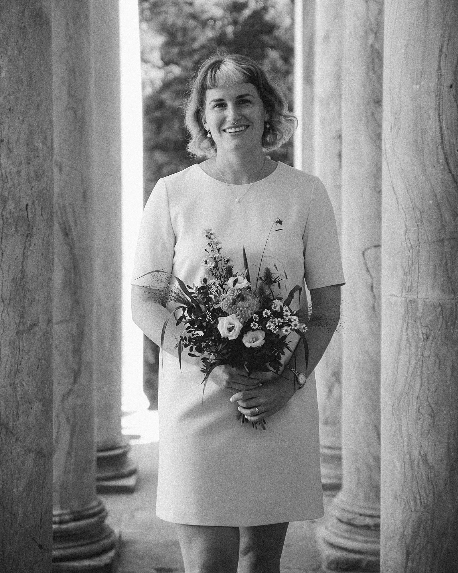 black and white portrait of a bride smiling at the camera, holding a wedding bouquet and wearing a short beige dress. She's standing in between rows of columns on both sides.