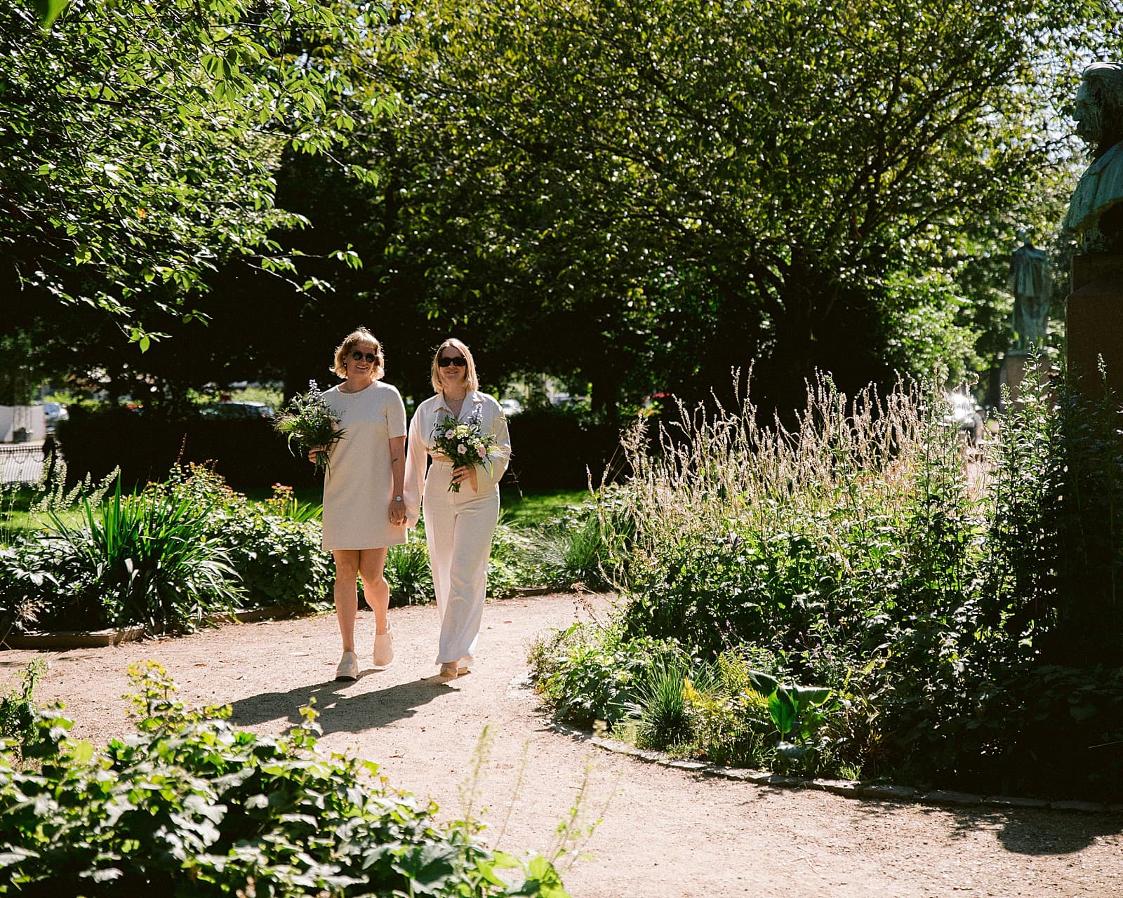 two brides wearing a beige jumpsuit and a beige dress walking towards the camera in a garden, holding a wedding bouquet