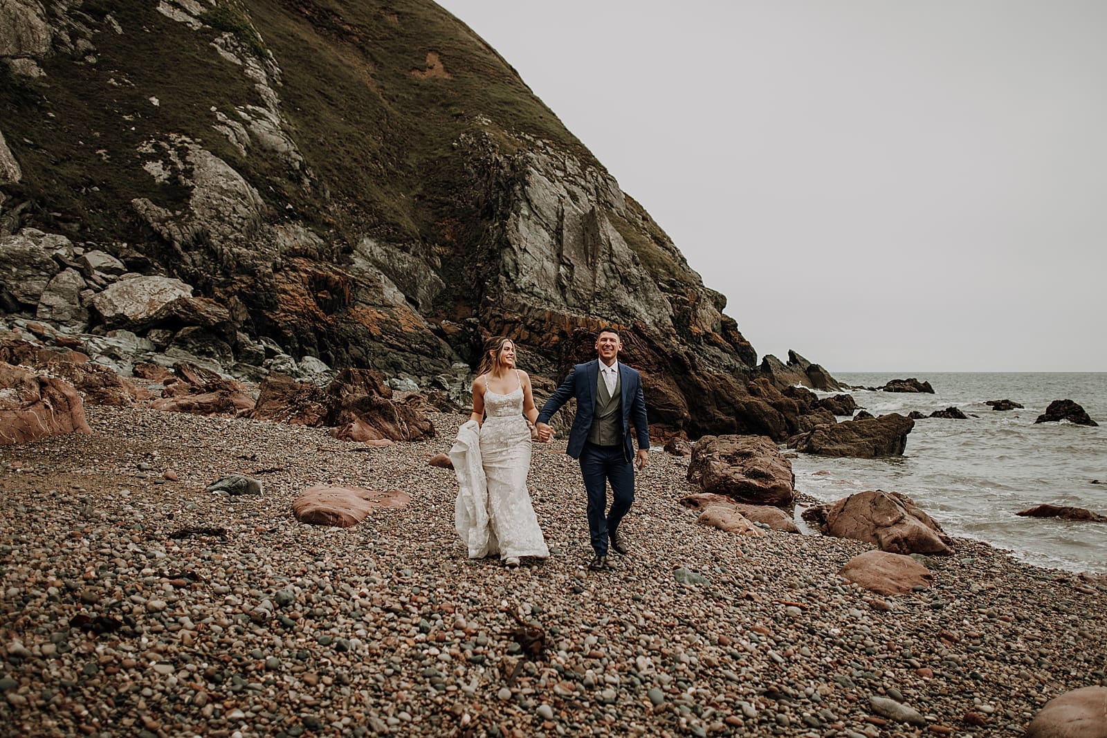 bride and groom are walking hand in hand along the beach in Ireland, smiling at each other and the camera