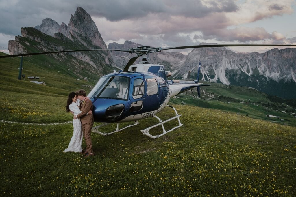 Bride and groom lean against the front of a blue helicopter that's parked on the Seceda mountain in the Dolomites in Italy
