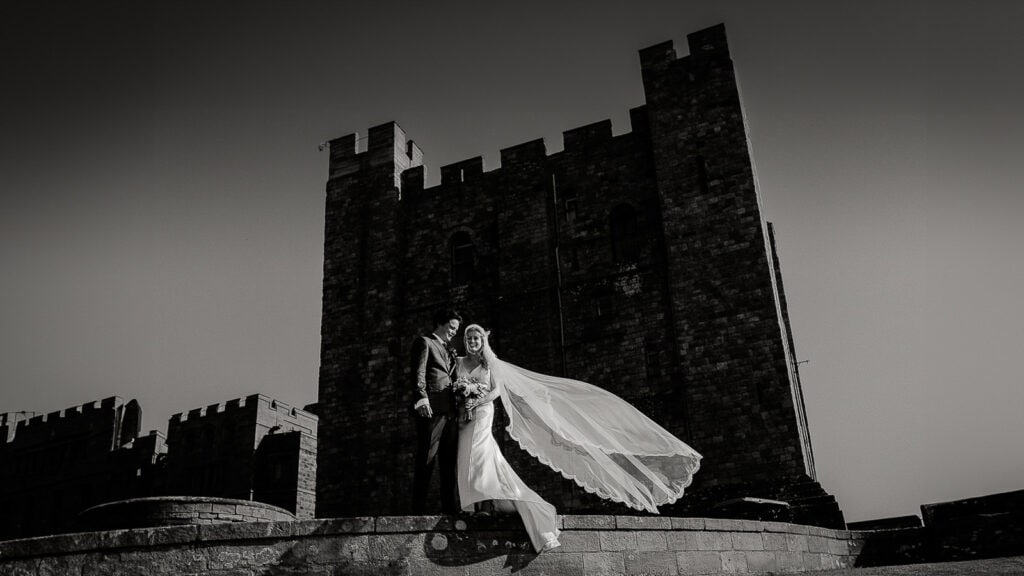 Bride and groom stand on the walls of a castle while her veil blows in the vind