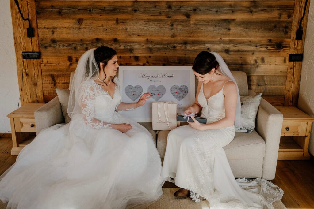 Two brides swap wedding day gifts for each other. They are sitting on a couch in a wooden ski chalet in Val Gardena in the Dolomites.