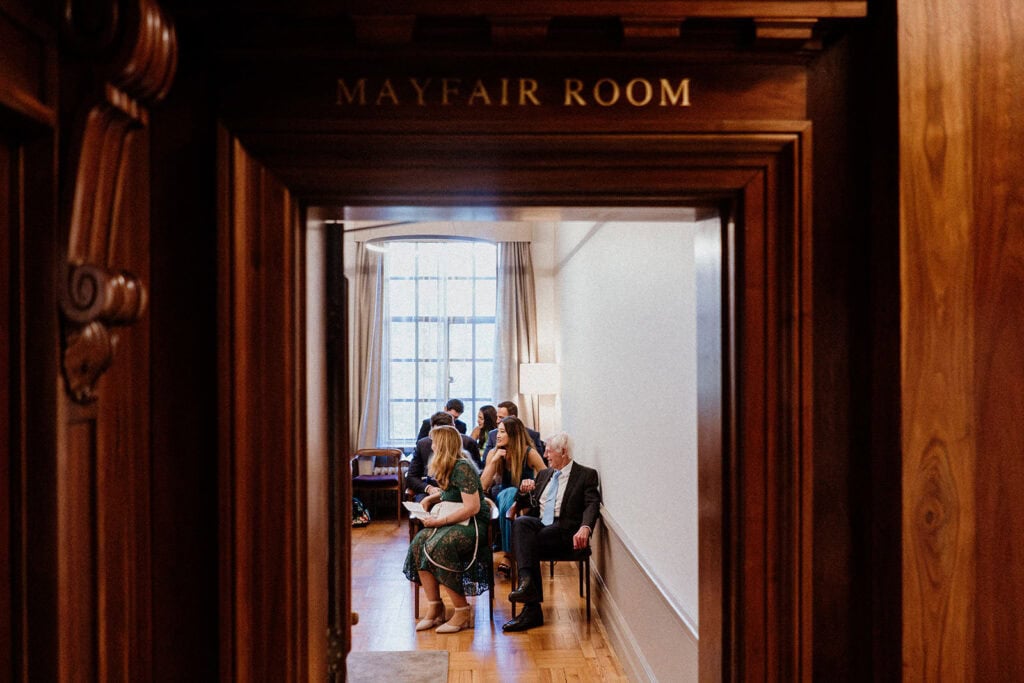 A photo looking through the door of the Mayfair room at wedding guests waiting for the ceremony at Old Marylebone Town Hall in London