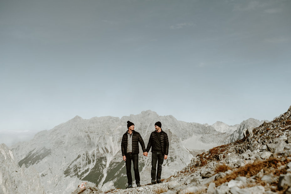 Two grooms hold hands. Both are wearing black suit pants and a black puffer jacket. There are mountains in the background behind them.
