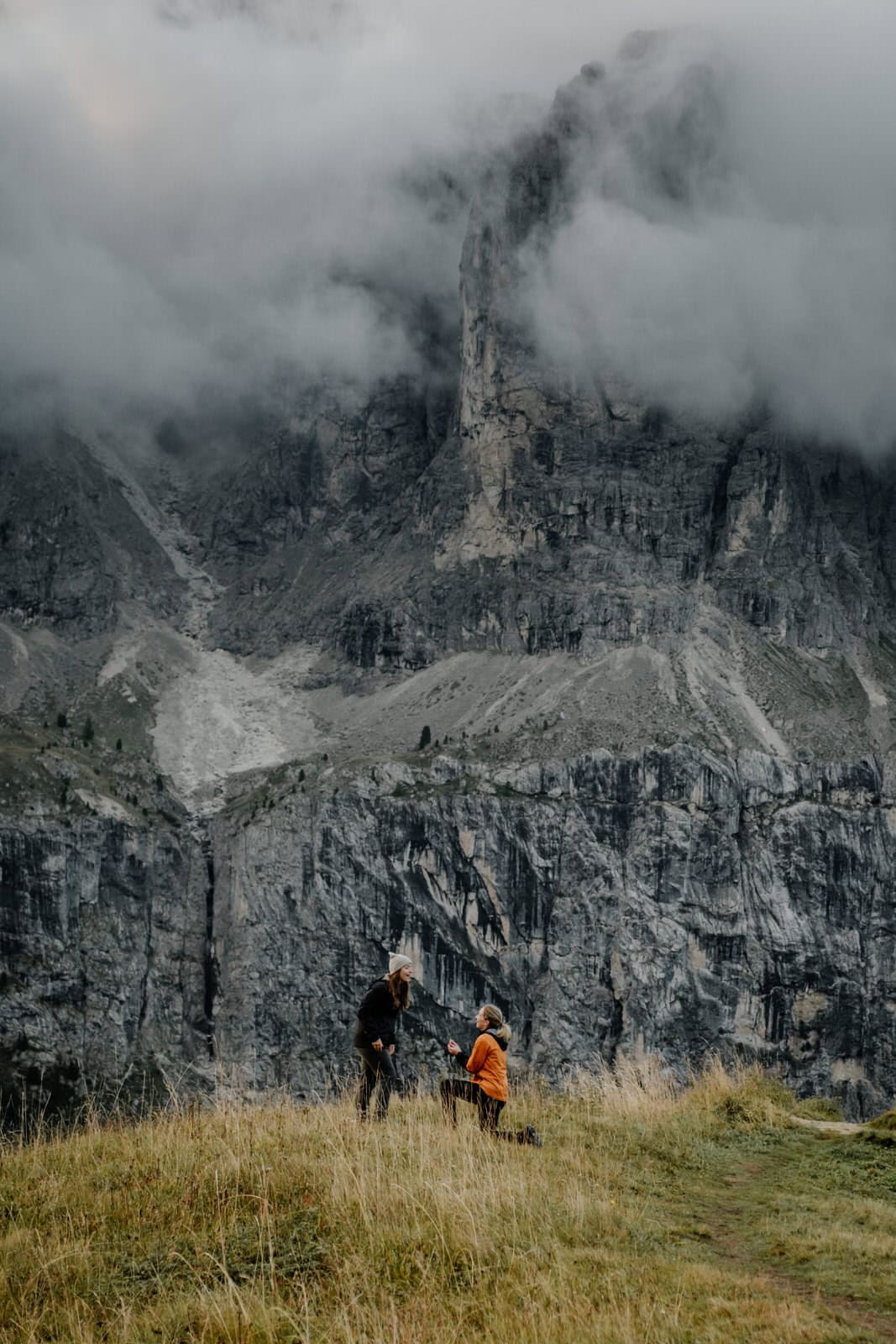 A photo of two women. One is kneeling down and proposing to the other. There are mountains behind them.