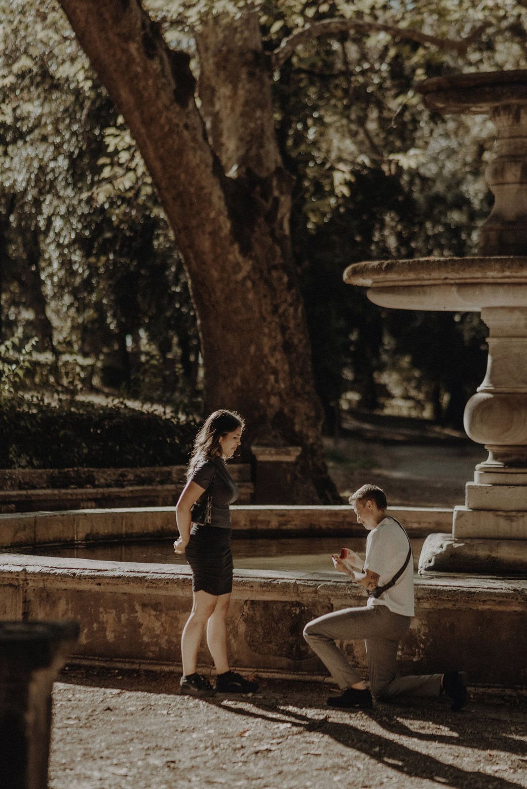 An LGBT proposal, one parter is down on one knee with a rung box in front of a fountain in Rome.