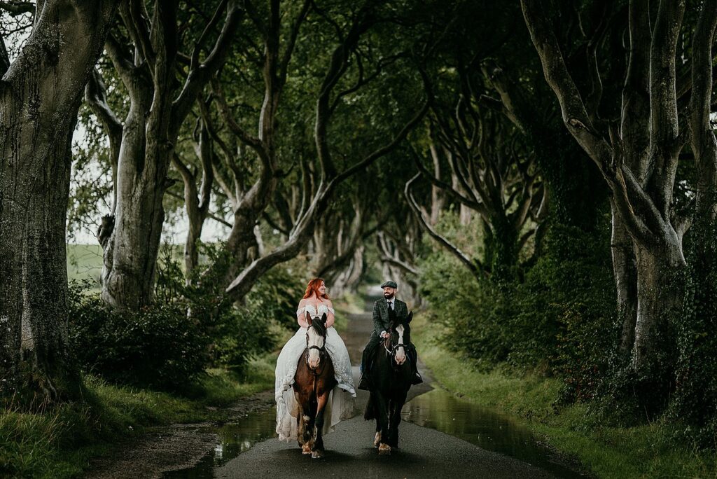 Image of a bride and groom riding horses down a road that is lined with old trees, known as the Dark Hedges in Northern Ireland